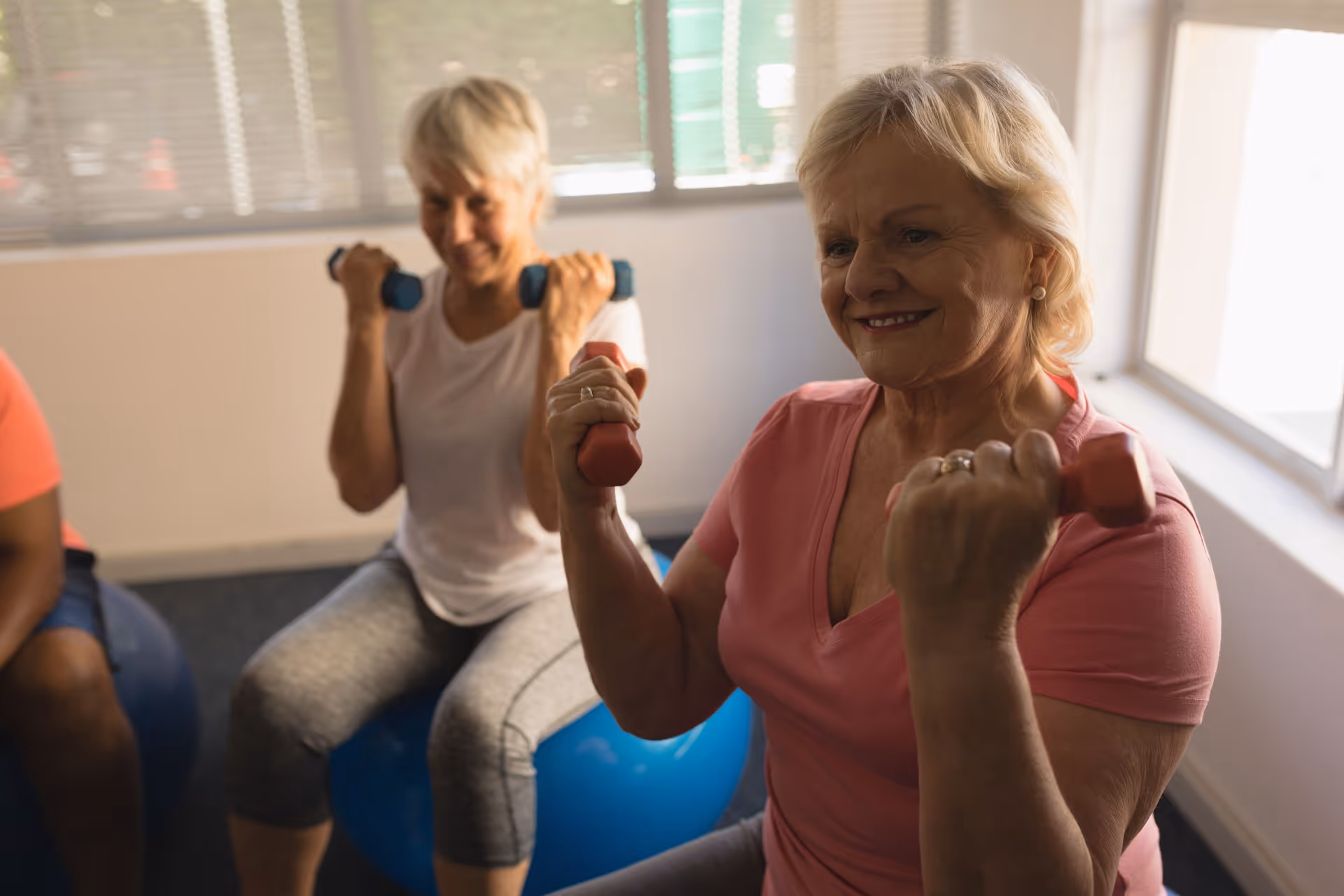 Two elderly women exercising indoors while sitting on blue exercise balls, each holding a pair of small dumbbells and smiling.