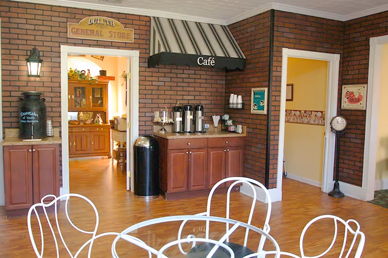 Interior view of a cozy café area with brick walls and wooden flooring. There is a coffee station with multiple coffee dispensers, cups, and condiments on a countertop beneath a black awning labeled 'Café'. To the left, there is an open doorway leading to a room labeled 'Duluth General Store' with wooden furniture. White metal chairs and a table are partially visible in the foreground.