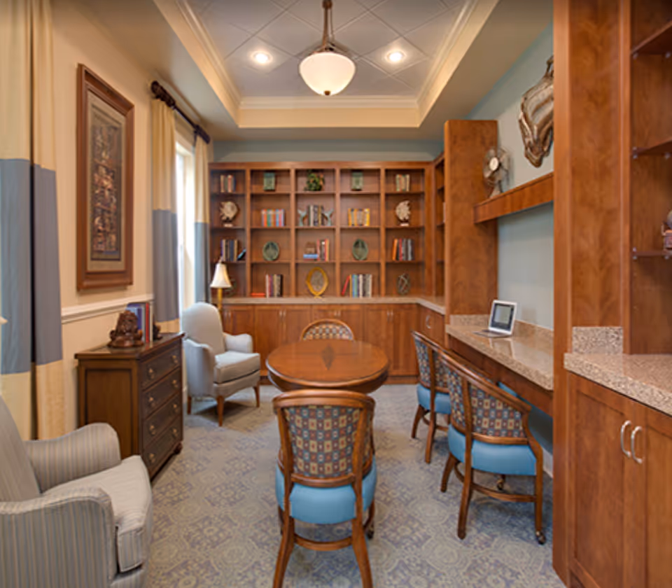 A cozy interior room with wooden bookshelves filled with books and decorative items, a round wooden table surrounded by four chairs with patterned upholstery, two armchairs, a small chest of drawers, and a countertop with a tablet on it. The room has a soft carpet, light blue walls, and a ceiling light fixture.