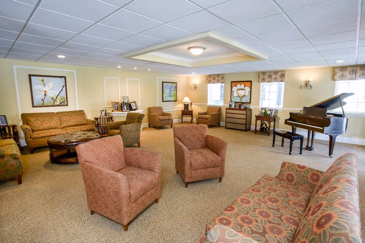 A spacious and well-lit living room with multiple patterned armchairs and sofas arranged around a central round ottoman. The room features beige walls adorned with framed floral artwork, several windows with patterned valances, a grand piano with a bench, and various small tables displaying framed photographs and decorative items.