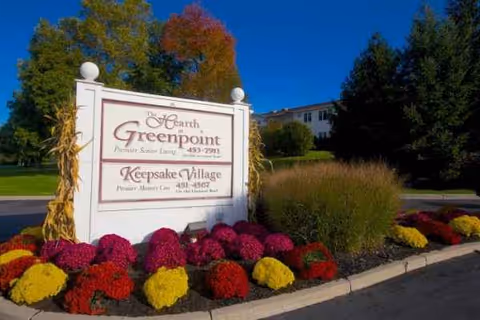 Outdoor sign for The Hearth at Greenpoint with colorful flower bed and building in background.