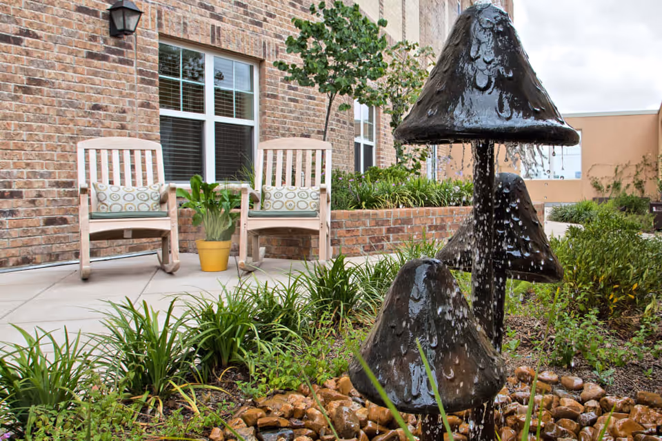 Outdoor patio area with two wooden chairs featuring patterned cushions, a yellow potted plant between them, and a decorative water fountain shaped like mushrooms surrounded by greenery and rocks in front of a brick building.