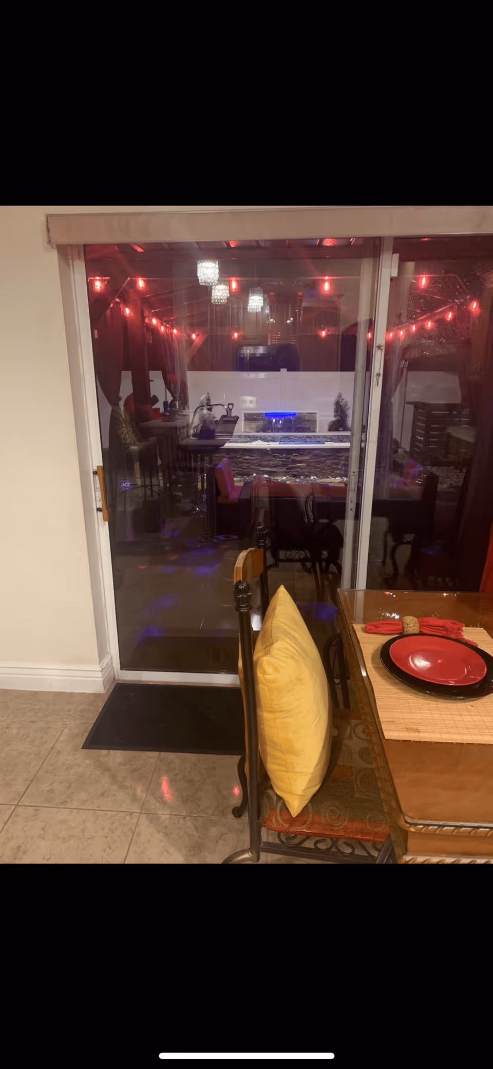 View of a dining area with a glass table set with red and black plates and a yellow cushion on a chair. Through a sliding glass door, an outdoor patio area is visible with red string lights, hanging lanterns, cushioned seating, and a water feature with blue lighting.