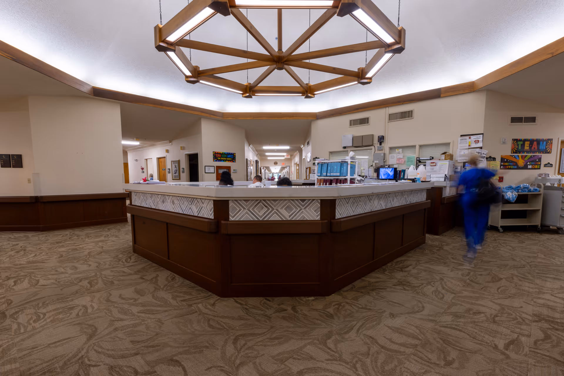 Interior view of a senior living facility reception or nurse station area with a large wooden counter in the center, beige walls, patterned carpet, and a geometric wooden light fixture on the ceiling. A person in blue scrubs is walking on the right side, and there are several people seated behind the counter. Various office supplies and informational posters are visible on the walls behind the counter.