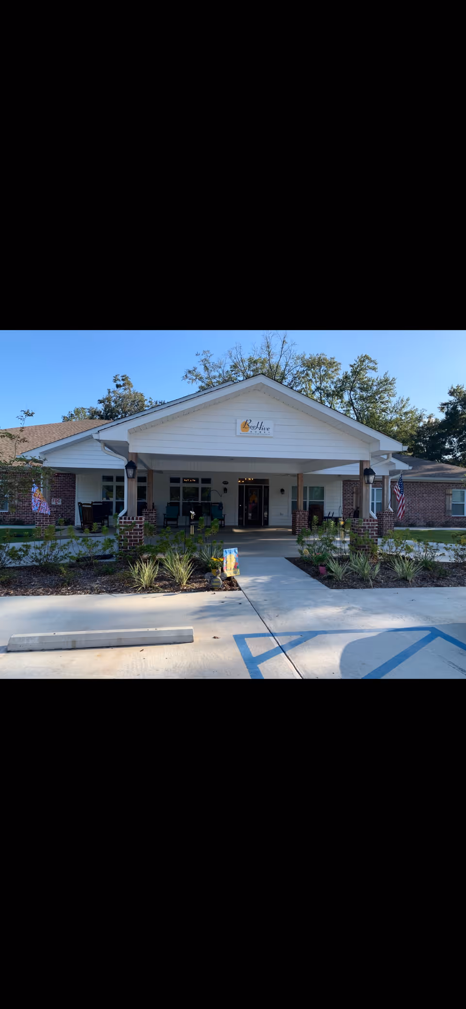 Front exterior view of BeeHive Homes Assisted Living facility with a covered entrance, brick columns, and a sign displaying the facility name. There are plants and shrubs along the walkway leading to the entrance, and American flags are visible on either side of the building.