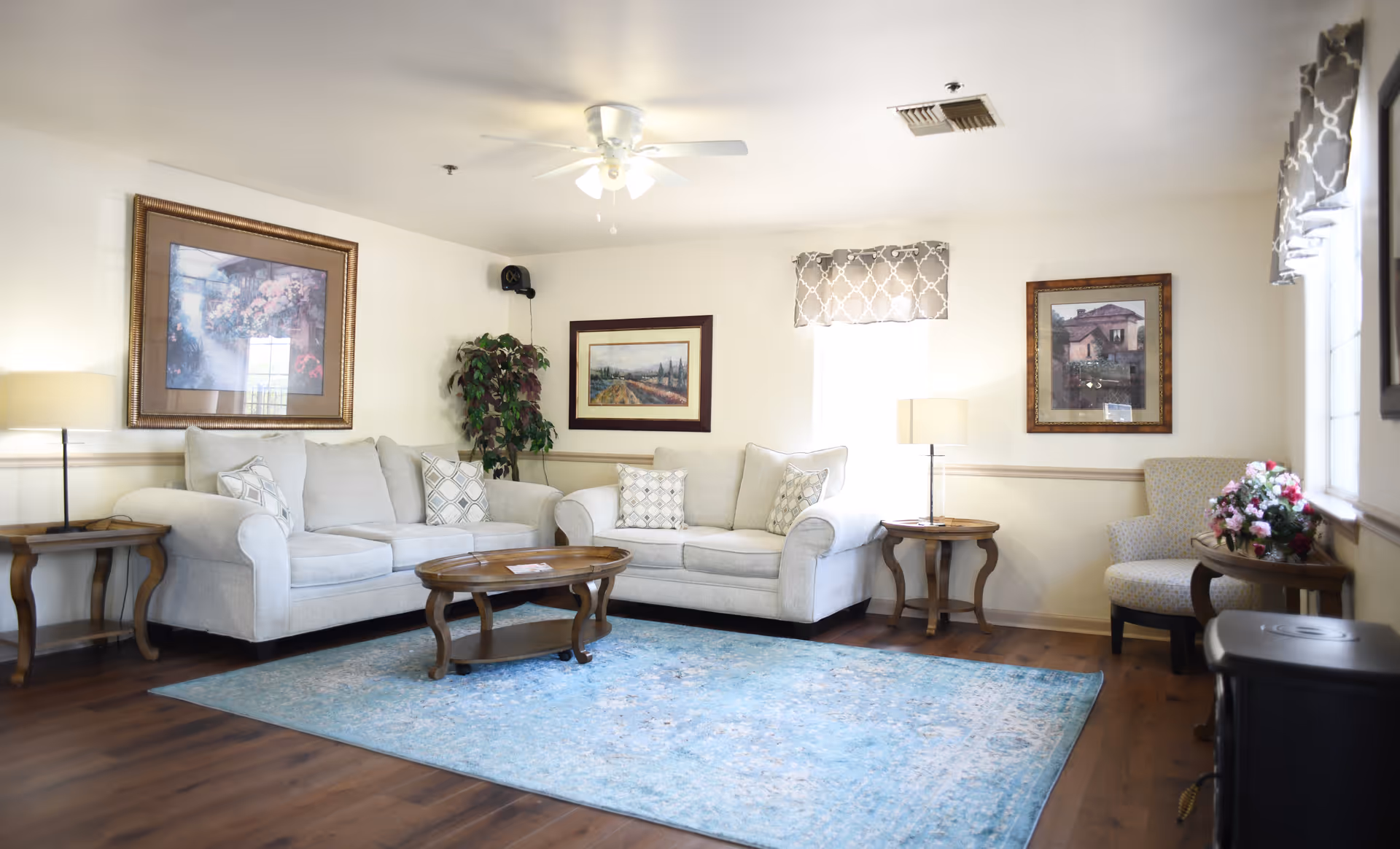Bright living room with light-gray sofas, a wooden coffee table on a blue area rug, framed artwork on the walls, and side tables with lamps.