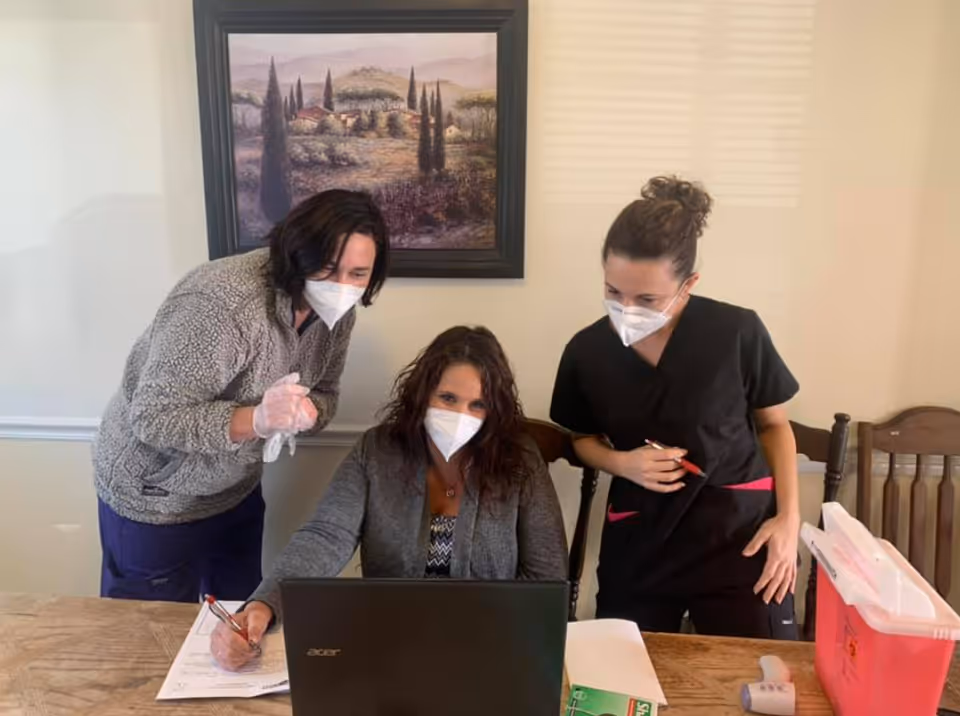 Three women wearing face masks are gathered around a table with a laptop and paperwork. One woman is seated and using the laptop, while the other two stand beside her looking at the screen. They appear to be in a meeting or collaborative work setting inside a room with a framed landscape painting on the wall behind them.