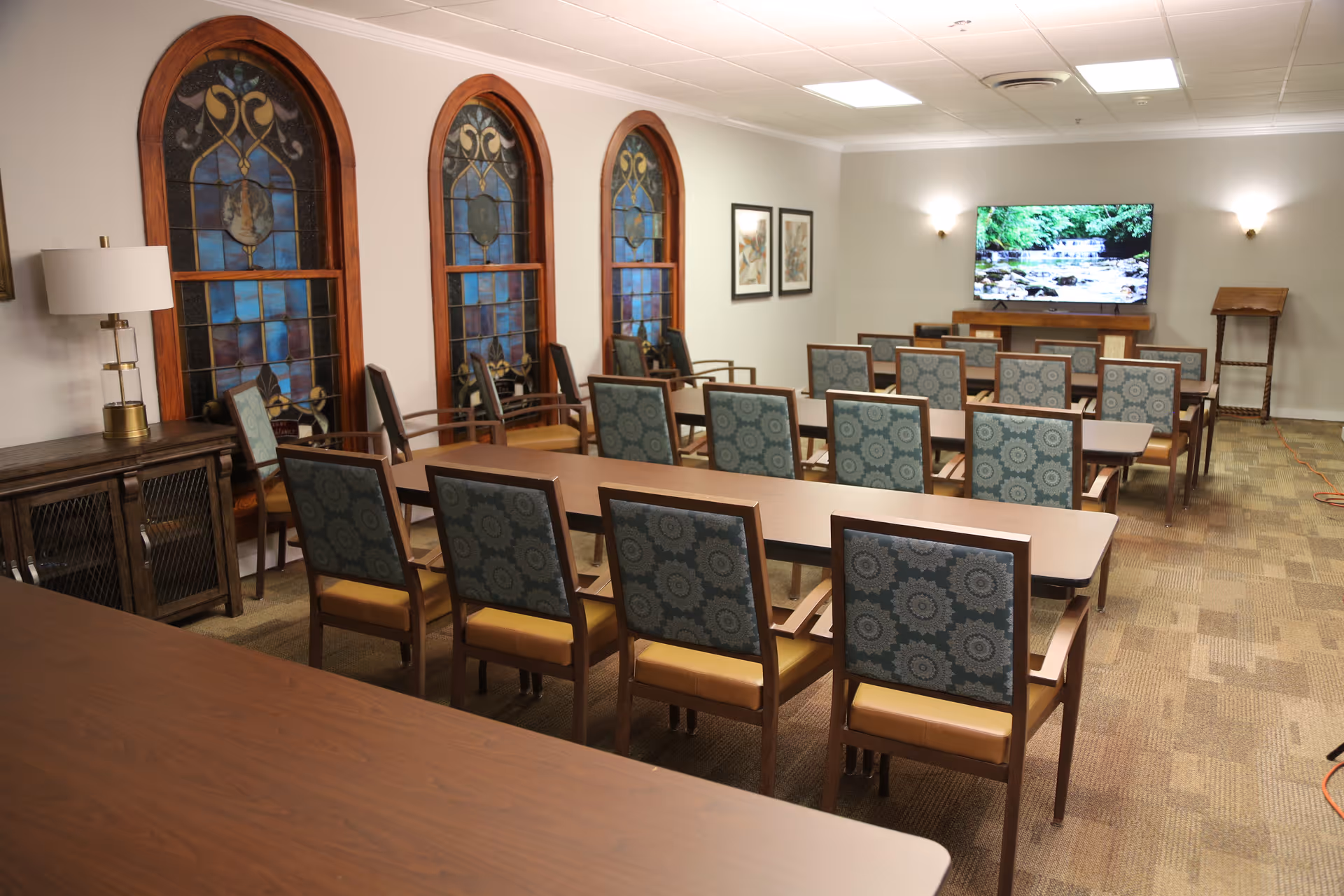 A room with multiple rows of tables and chairs arranged facing a wall-mounted television displaying a nature scene. The room features three large stained glass windows on one side, a sideboard with a lamp, framed artwork on the walls, and carpeted flooring.