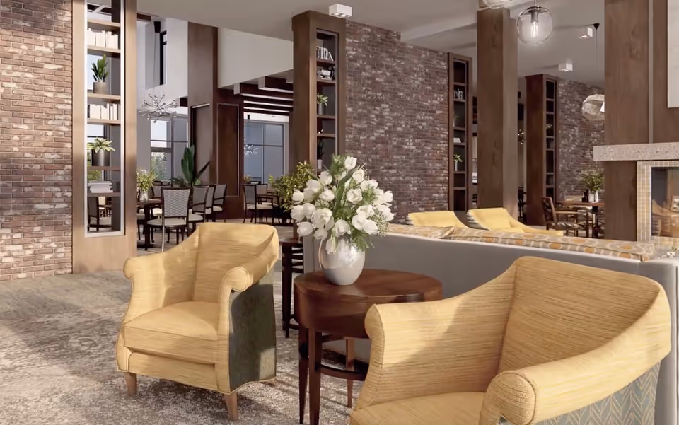 Open communal lounge with yellow armchairs around a round table holding a vase of white flowers, brick walls and dining tables visible in the background.