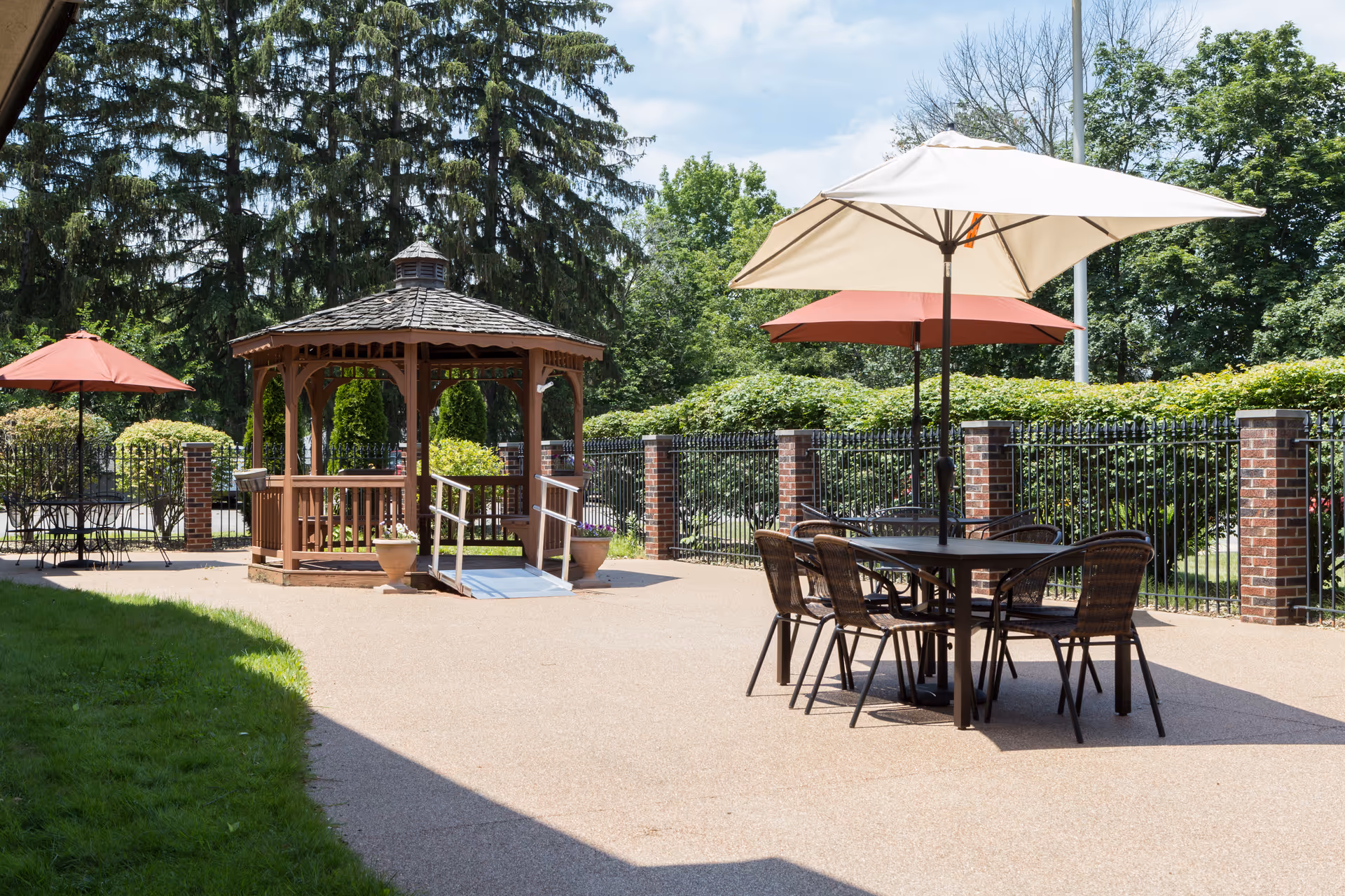 Outdoor courtyard featuring a wooden gazebo, patio tables with umbrellas, and a fenced garden area.