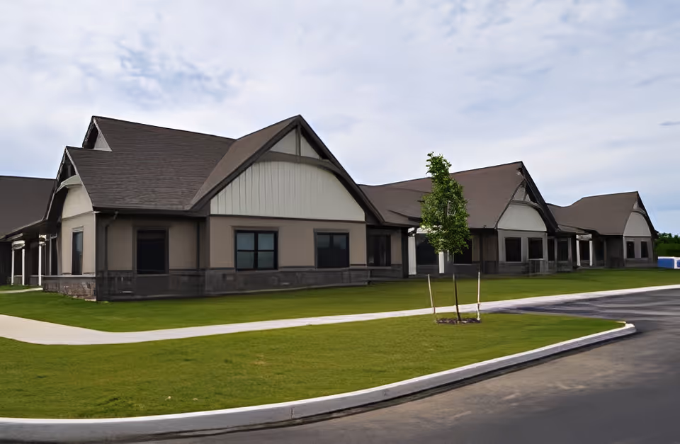 Exterior view of a single-story building with multiple peaked roofs, beige and brown walls, and several windows. The building is surrounded by a well-maintained green lawn, a paved walkway, and a paved driveway. A small tree is planted near the walkway.