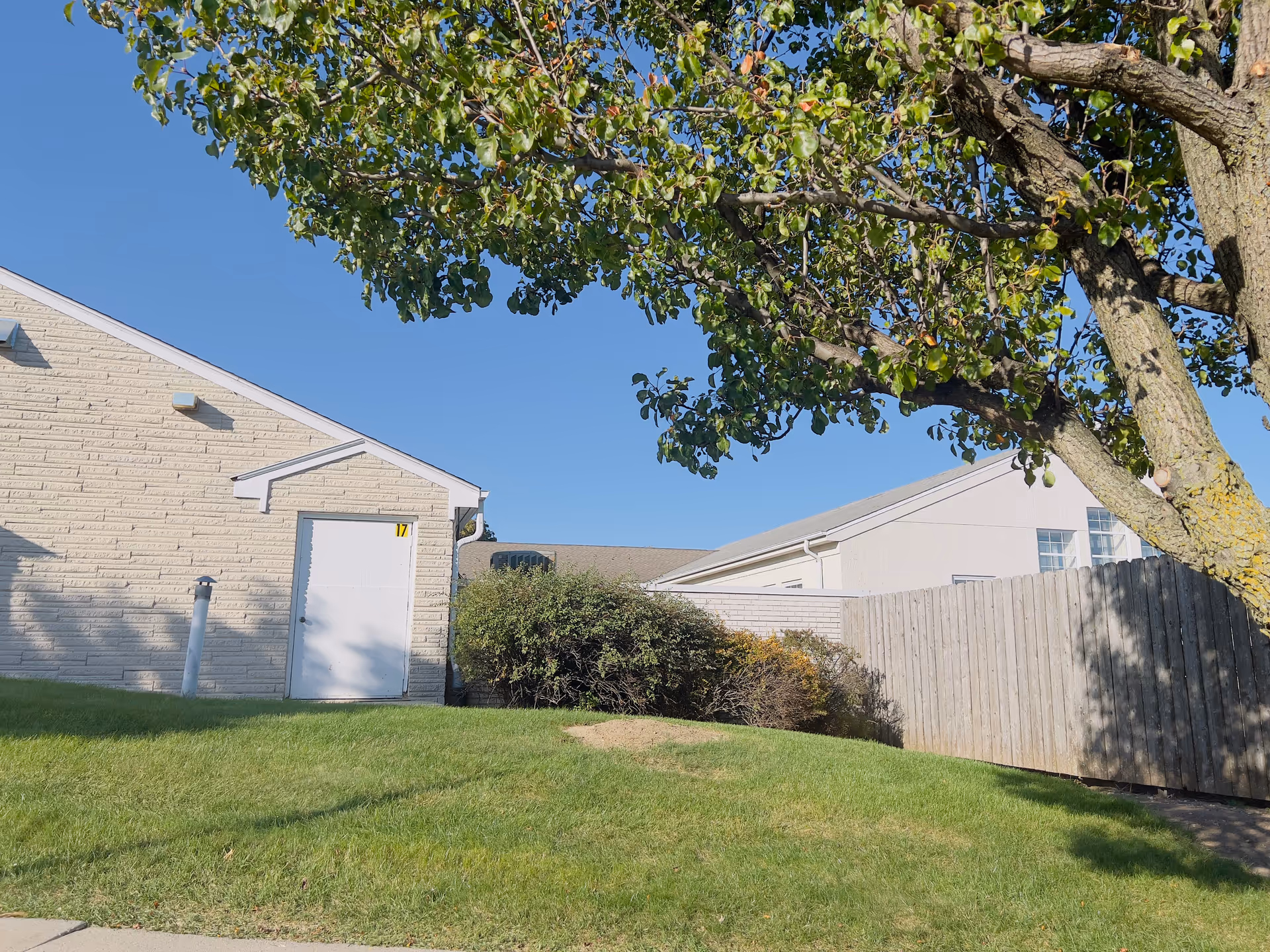 Exterior view of a building at Franciscan Gardens with a white door marked with the number 17, surrounded by green grass, bushes, a wooden fence, and a large tree under a clear blue sky.