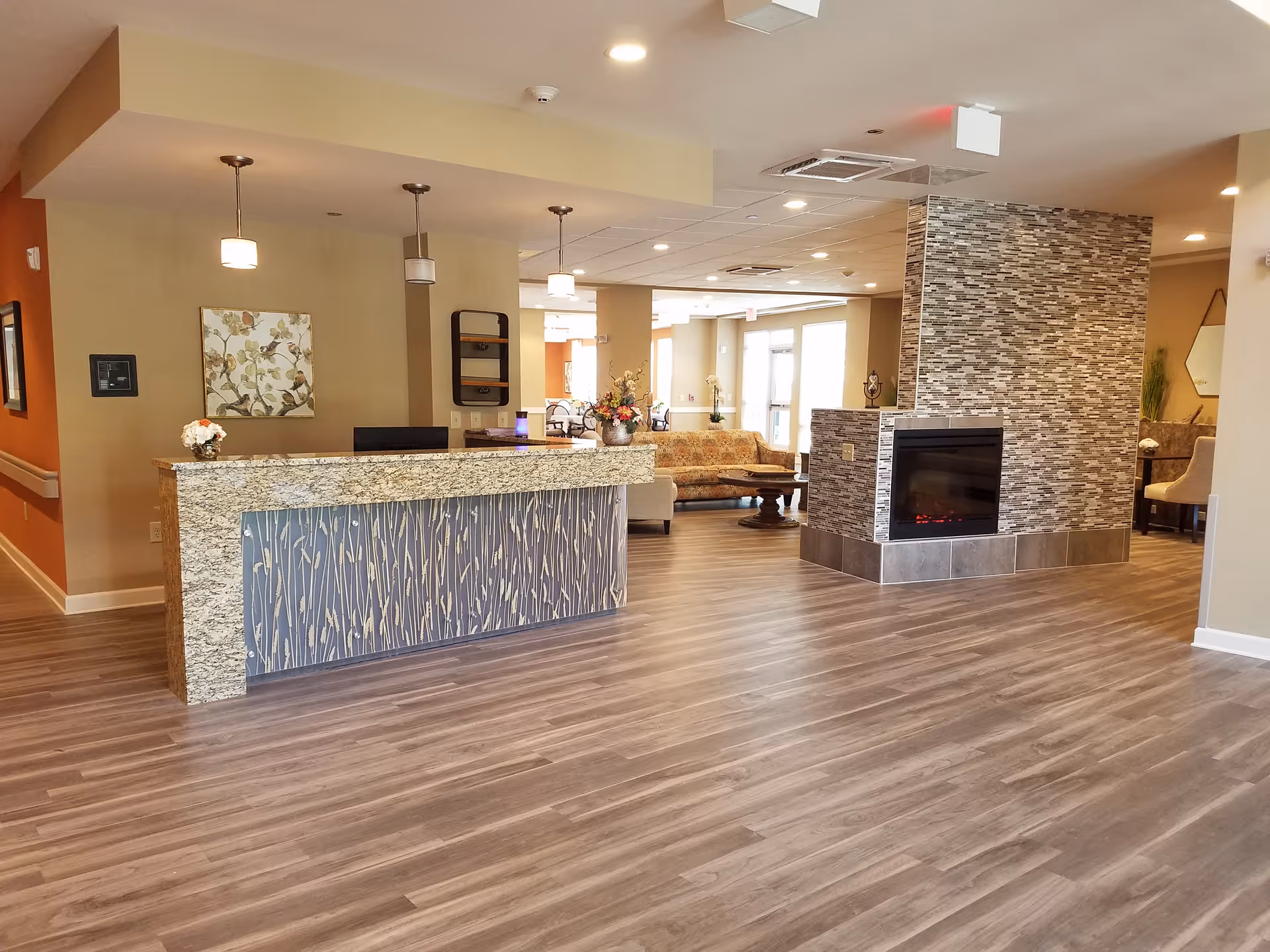 A spacious and modern senior living facility lobby with a granite reception desk featuring a decorative front panel. The area includes a cozy seating arrangement with a floral-patterned sofa and armchairs around a wooden coffee table. A contemporary double-sided fireplace with a stone tile surround divides the space. The room has wood-look flooring, warm beige walls, and ceiling lights providing bright illumination.