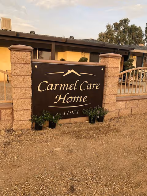 Outdoor view of the entrance sign for Carmel Care Home, mounted on a stone wall with a metal fence and a building in the background under a partly cloudy sky.