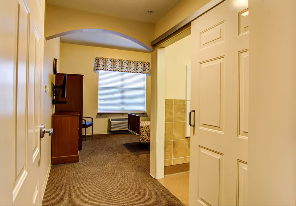 View into a senior bedroom showing a partially open door, TV cabinet, window with a valance, and a bed with an air conditioning unit below the window.