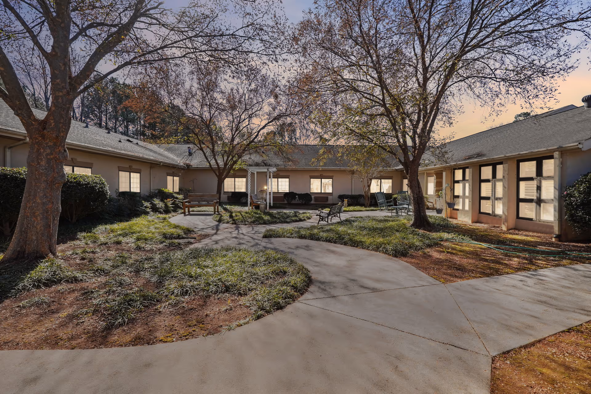 Outdoor courtyard area at a senior living facility with a paved walkway, benches, trees with sparse leaves, and surrounding single-story building with multiple windows.