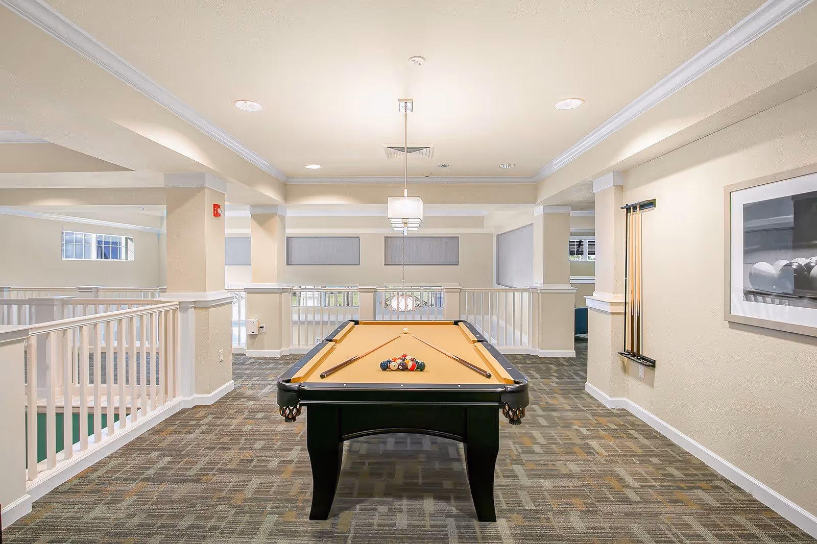 Interior view of a recreational room with a pool table in the center. The pool table is set up with balls and two cues resting on it. The room has beige walls, carpeted floor, white railings, and ceiling lights. Pool cues are mounted on the wall to the right, and a framed picture hangs nearby.