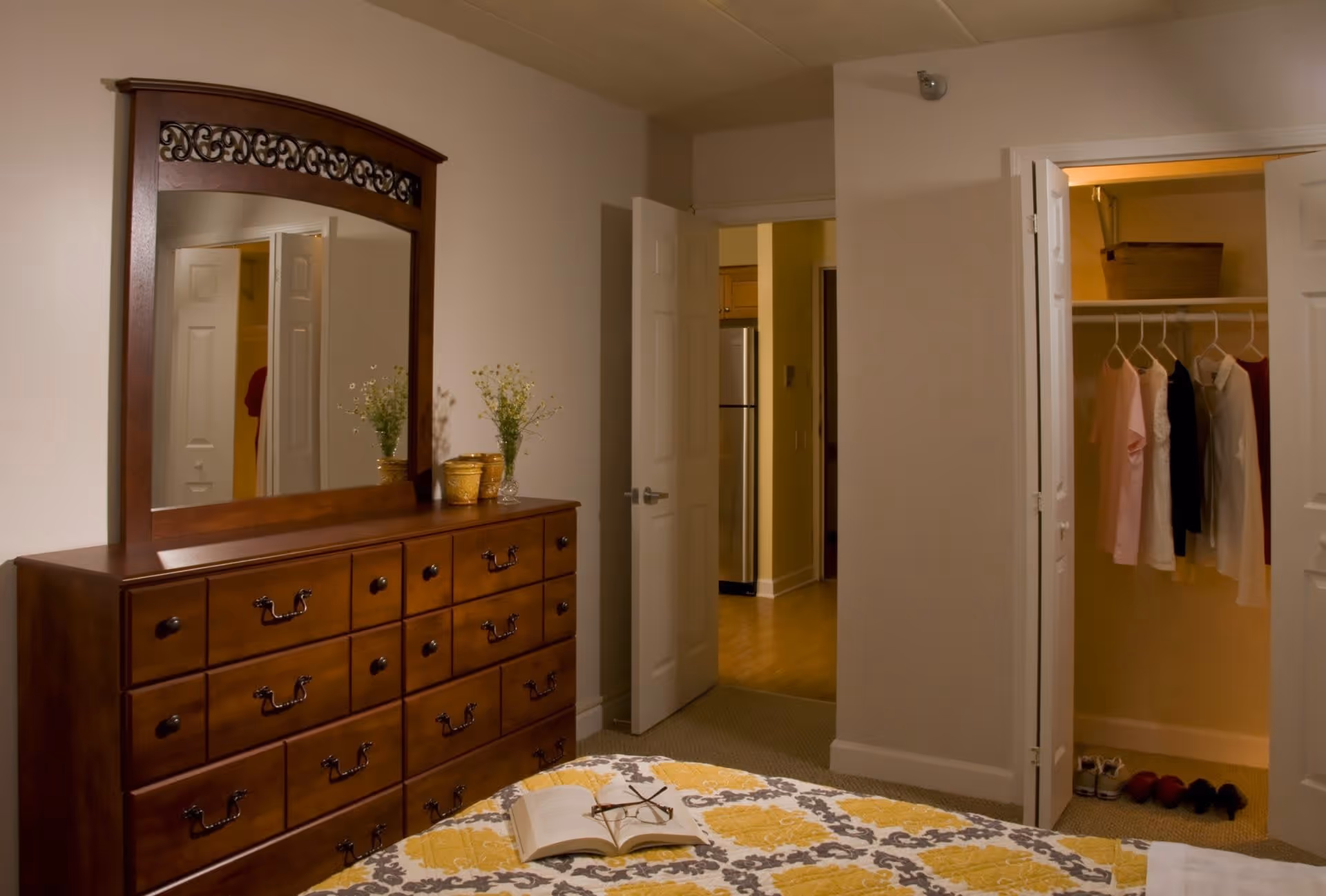 A bedroom with a wooden dresser topped with a large mirror and two small vases with flowers. The bed has a patterned yellow, gray, and white quilt with an open book and eyeglasses on it. There are two open doors, one leading to a hallway with a refrigerator visible, and the other to a closet with hanging clothes and shoes on the floor.