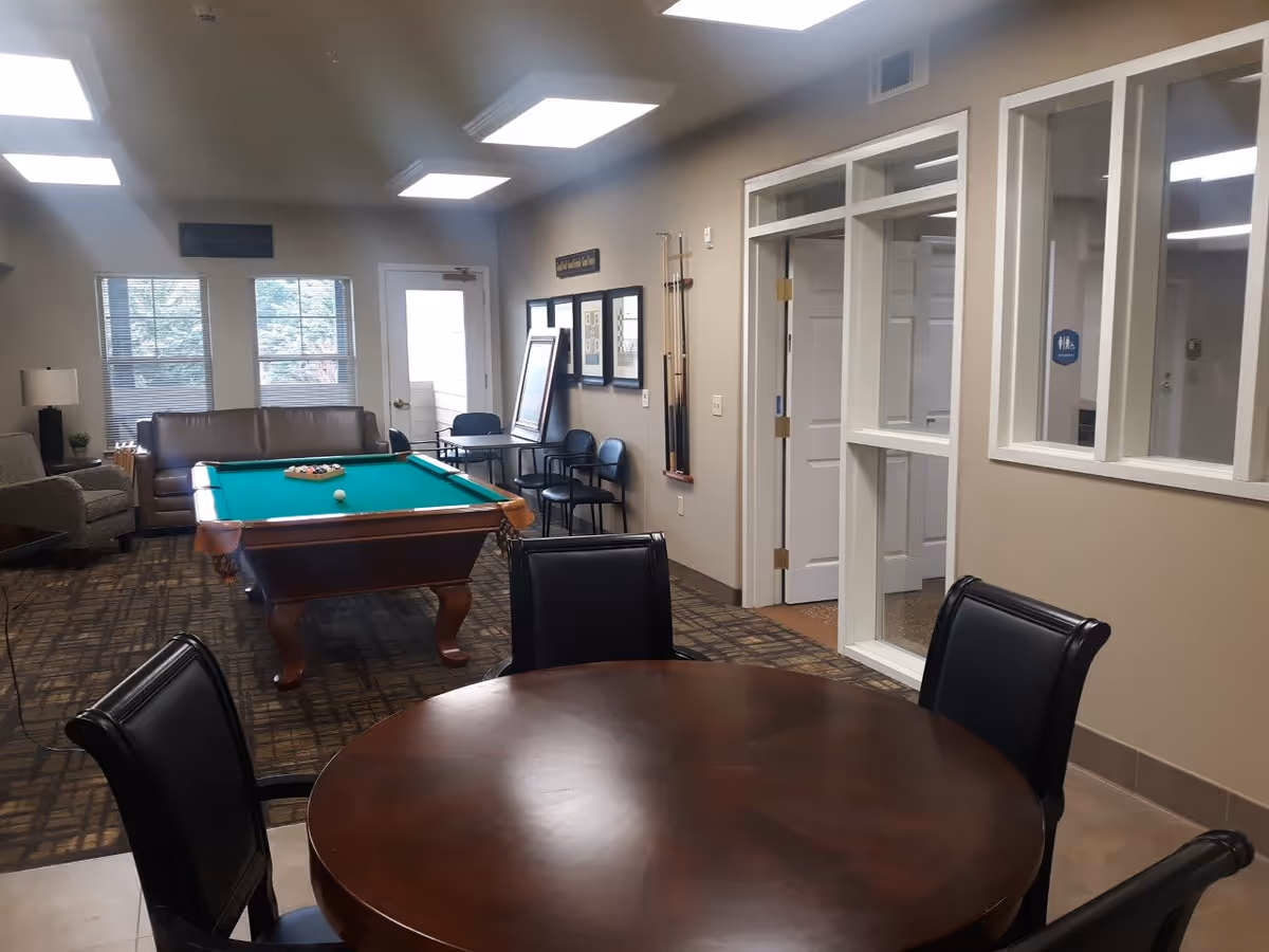 Interior room with a pool table in the center, a brown leather sofa and an armchair near two windows, a round wooden table with four black chairs in the foreground, and a wall with windows and doors on the right side.