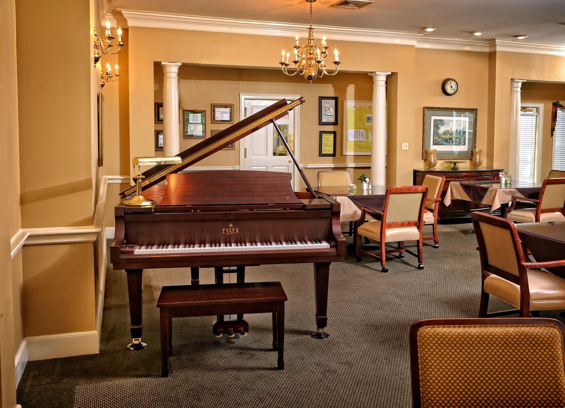 A dining room/lounge with a grand piano in the foreground, tables and chairs, chandeliers, and framed artwork on the walls.