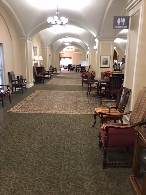A long hallway in a senior living facility with arched ceilings and chandeliers. The hallway is carpeted with patterned rugs and lined with various wooden chairs and small tables. There is a sign indicating the location of the restroom on the right side of the hallway.
