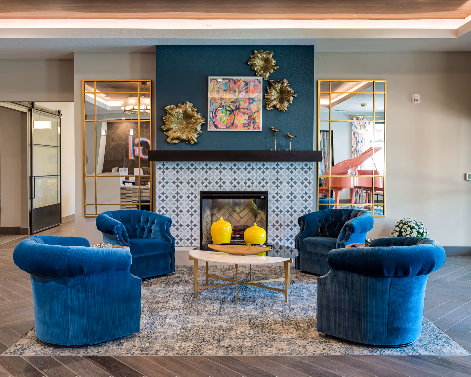 A cozy seating area with four blue velvet armchairs arranged around a round coffee table with a marble top and gold legs. Behind the seating area is a decorative fireplace with patterned tiles and a dark mantel. Above the mantel are gold floral wall decorations and a colorful abstract painting. Two large gold-framed mirrors flank the fireplace, reflecting parts of the room including a pink grand piano. The floor has a patterned rug and wood-like tiles.
