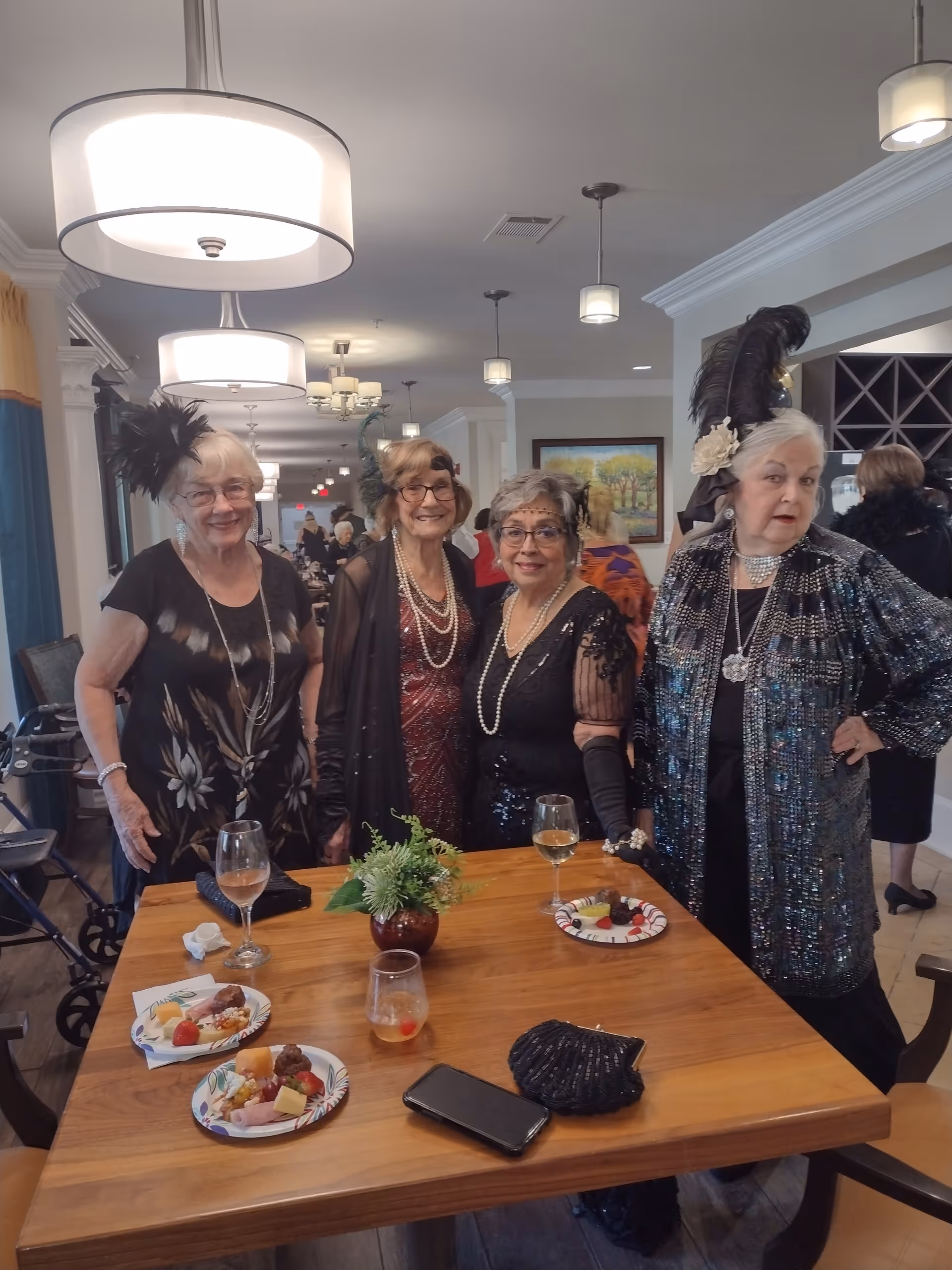 Four women in vintage, flapper-style outfits stand behind a wooden table with plates, drinks, and a small plant in a well-lit communal dining area.