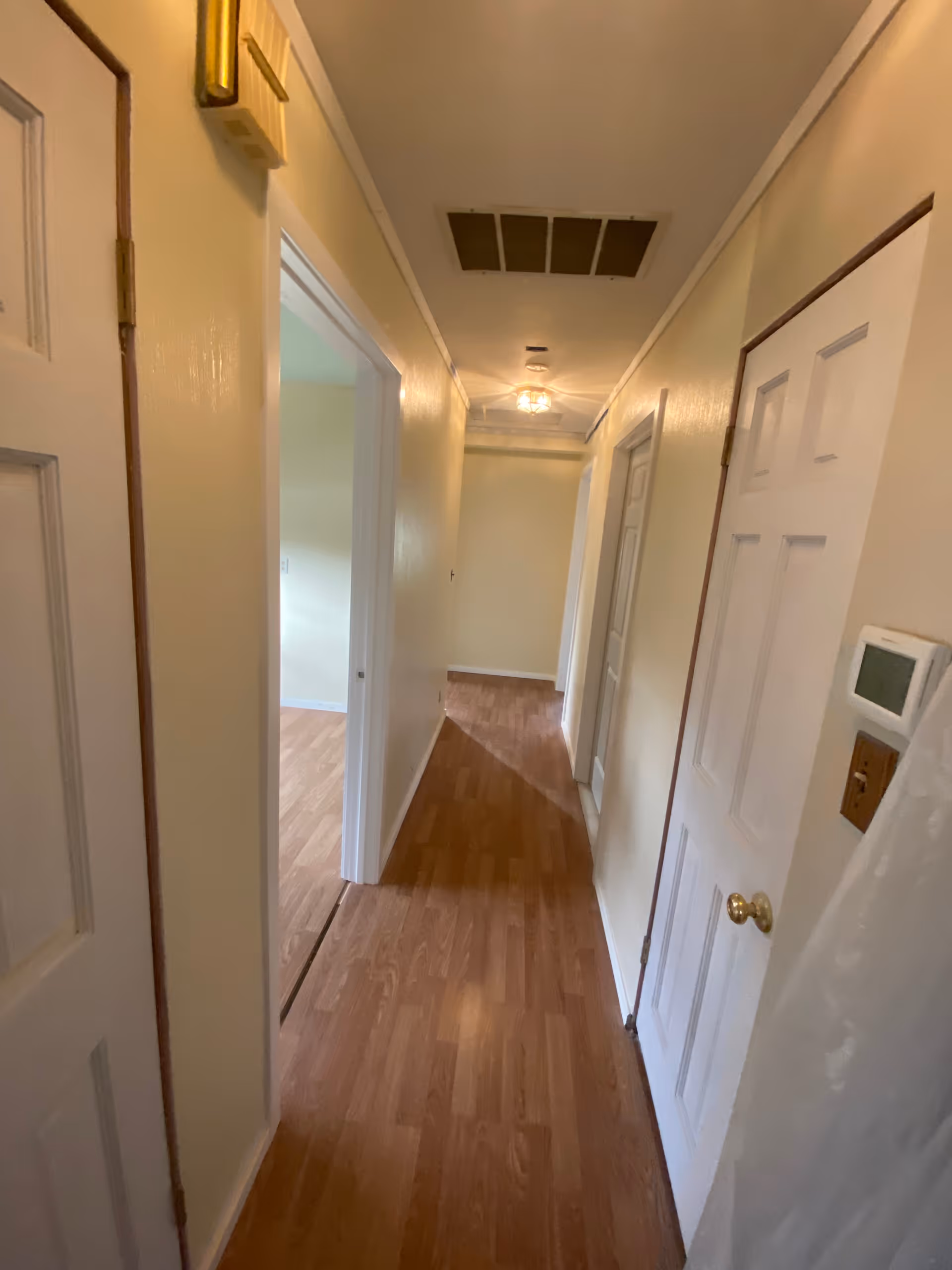 Narrow hallway with wood laminate flooring, cream-colored walls, white doors on both sides, and a ceiling vent and light fixture.