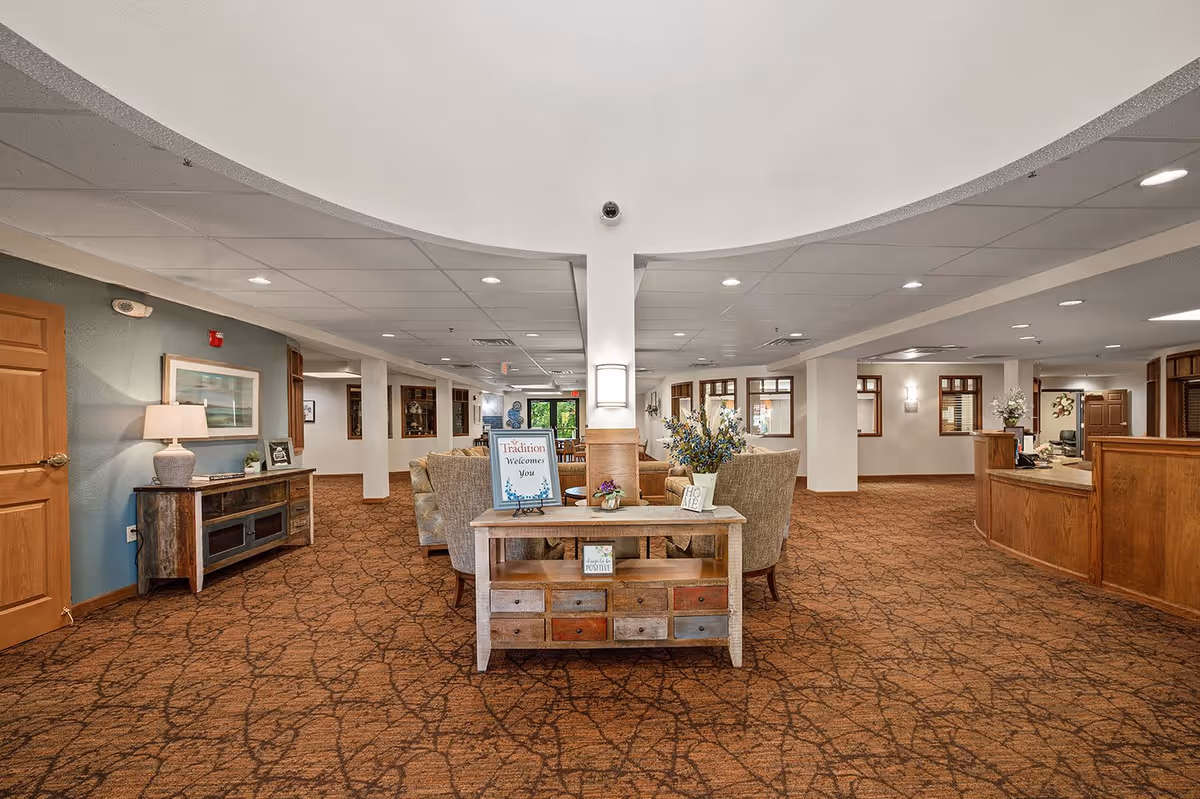Spacious and warmly decorated interior of a senior living facility lobby with patterned brown carpet, a wooden console table with flowers and a welcome sign that reads 'Tradition Welcomes You', comfortable seating area with armchairs, wooden reception desk on the right, and soft lighting from ceiling fixtures and wall sconces.