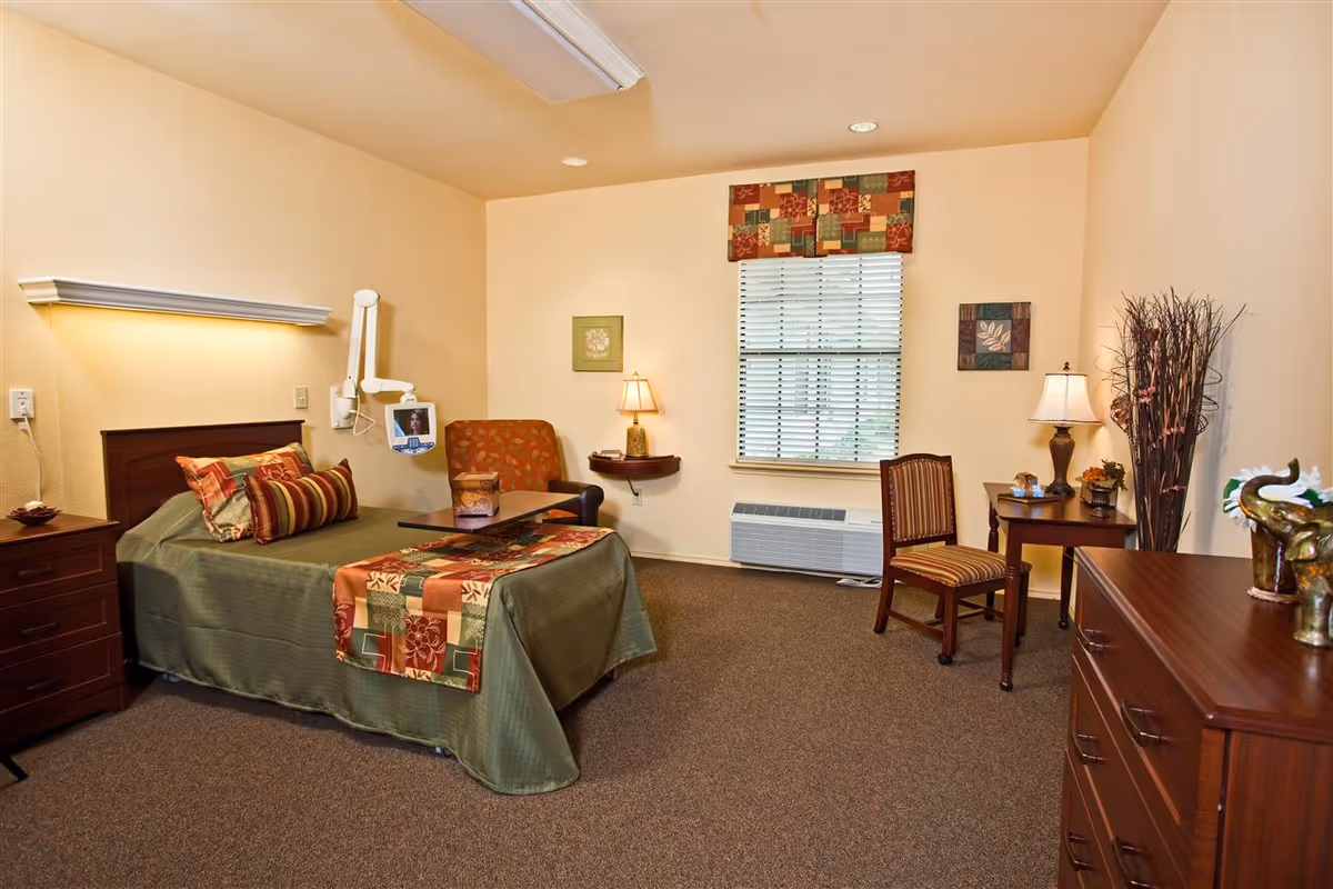 Well-lit senior living bedroom with a single bed, bedside tables, chair, dresser, and a window with blinds.