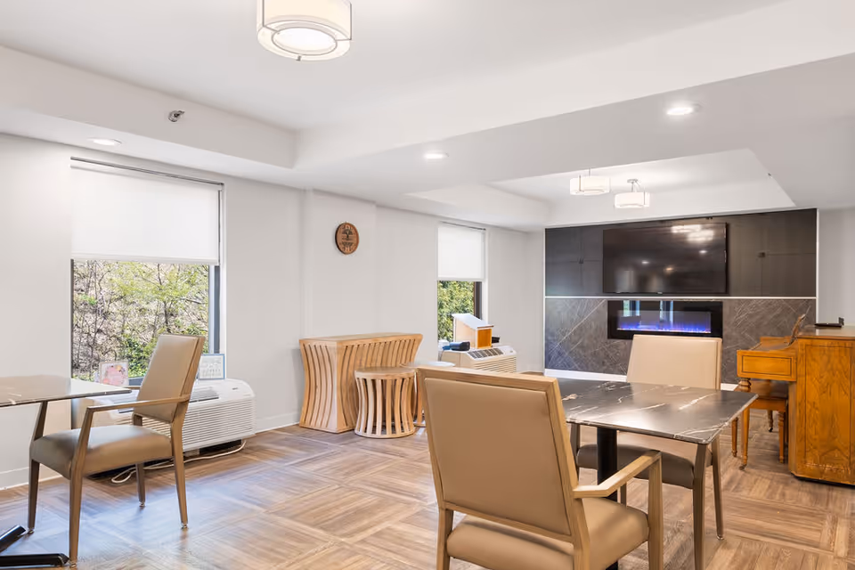 Bright common area with tables and chairs, a wall-mounted TV above a modern fireplace, and windows overlooking greenery.