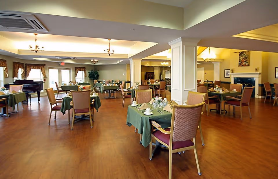 A spacious dining room in a senior living facility with multiple tables covered in green tablecloths, each set with cups, napkins, and silverware. The room features wooden flooring, comfortable chairs with red cushions, chandeliers, a fireplace, and large windows with curtains allowing natural light to enter.