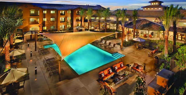 Evening view of an outdoor pool area at a senior living facility with lounge chairs, shaded seating areas, palm trees, and surrounding buildings with warm lighting.