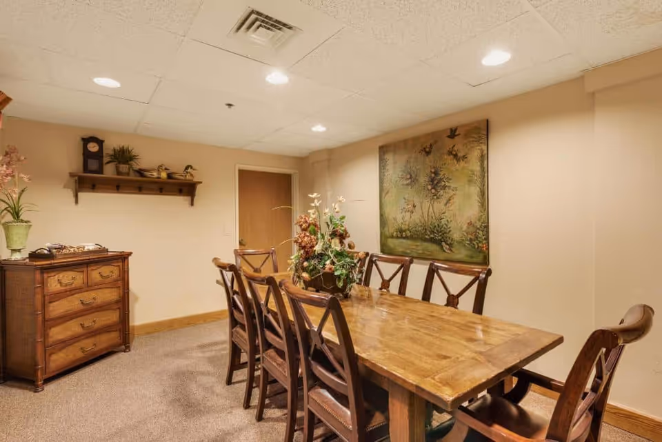 A wooden dining table with multiple chairs and a floral centerpiece in a warmly lit room with wall art and a chest of drawers.