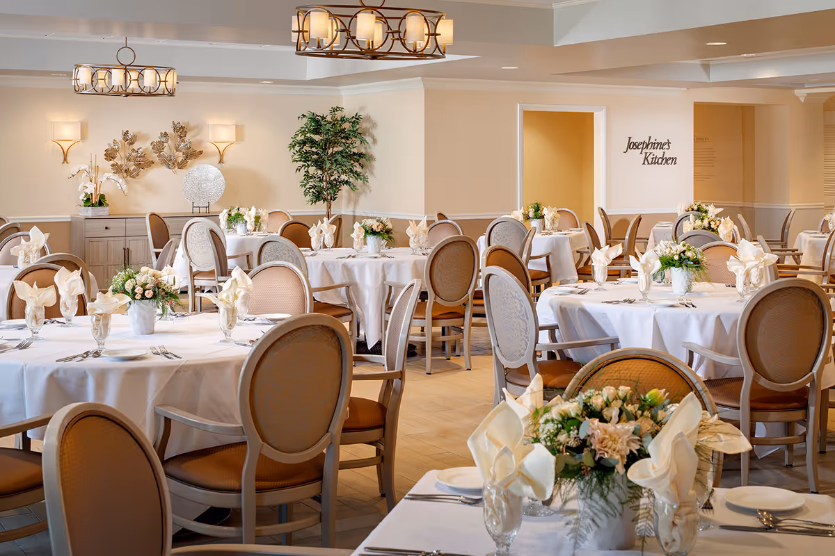 A bright and elegant dining room with round tables covered in white tablecloths, each set with folded napkins, glassware, and floral centerpieces. The room features beige walls, decorative wall art, and two modern chandeliers hanging from the ceiling. A sign on the wall reads 'Josephine's Kitchen'.