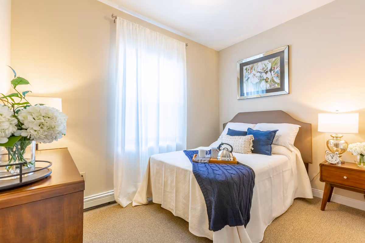 A cozy bedroom with a neatly made bed featuring white bedding and blue accent pillows. A blue knitted throw is draped across the foot of the bed with a wooden tray holding a kettle and a cup. The room has beige walls, a window with sheer white curtains, a wooden dresser with a vase of white flowers, and a bedside table with a lamp, clock, and another small vase of white flowers. A framed floral painting hangs above the bed.
