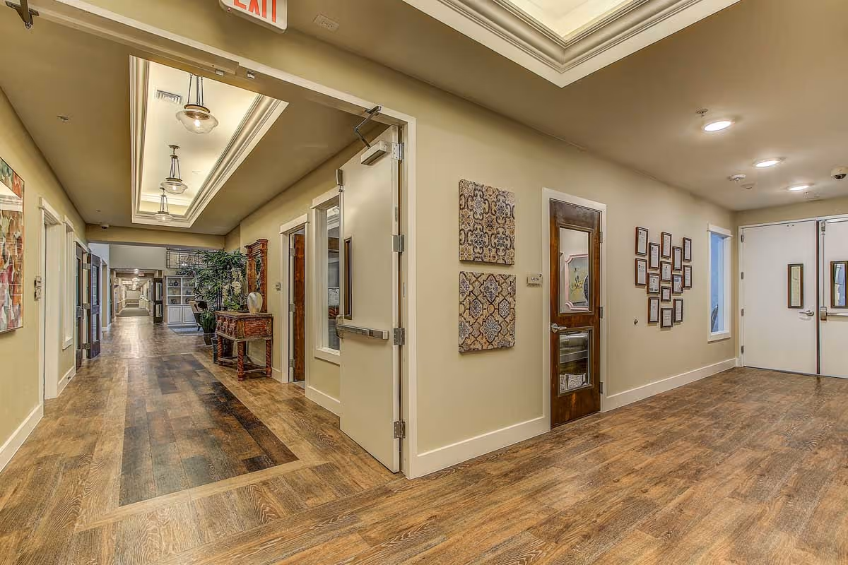 Interior hallway of a senior living facility with wood flooring, beige walls, decorative wall art, and ceiling lights. There is a wooden table with plants and vases along the hallway, and several doors leading to different rooms. An exit sign is visible above one door.