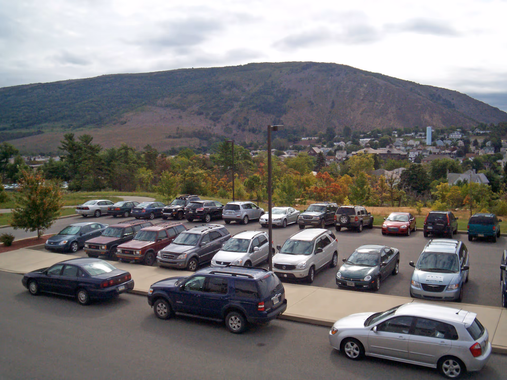 A parking lot filled with various cars in front of a scenic view of a large hill and a small town with houses and trees under a cloudy sky.