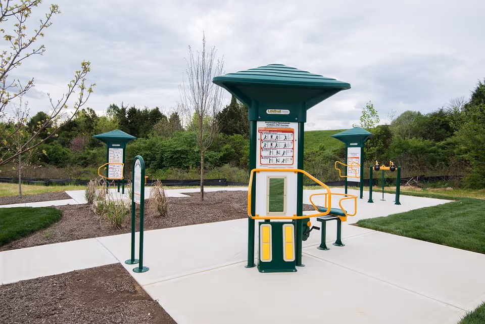 Outdoor exercise equipment stations with instructional signs on a paved area surrounded by grass and trees under a cloudy sky.