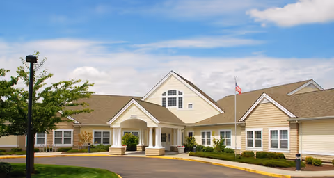Beige single-story senior living building with a covered entrance, flagpole, and landscaped grounds under a blue sky.