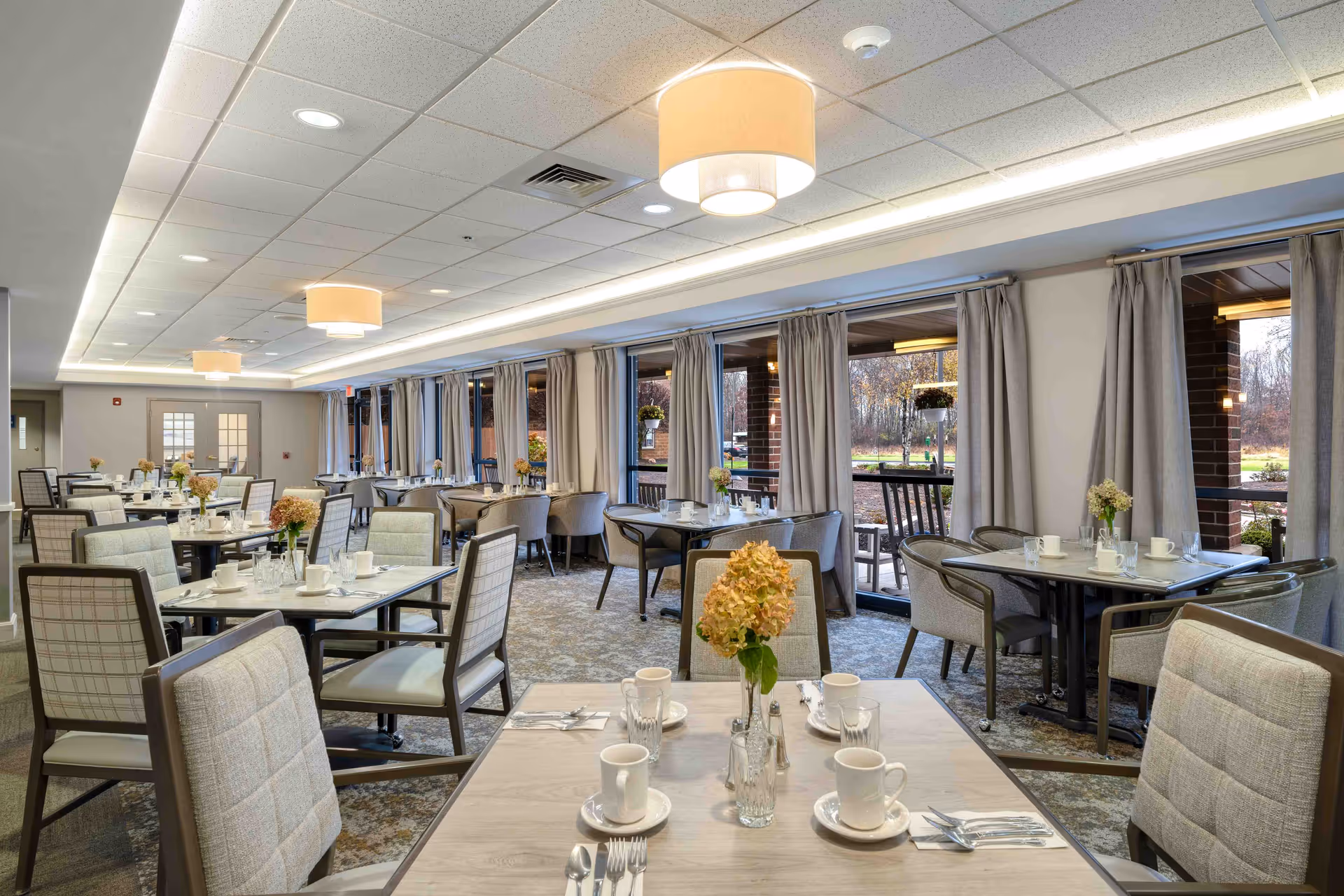 A bright and spacious dining room in a senior living community with multiple tables set with white cups, plates, silverware, and vases holding flowers. Large windows with beige curtains line one side of the room, allowing natural light to enter and providing a view of an outdoor patio area with rocking chairs. The ceiling has recessed lighting and modern hanging light fixtures.