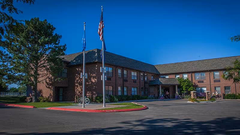 Two-story brick senior living building with flagpoles and a circular driveway beneath a clear blue sky.