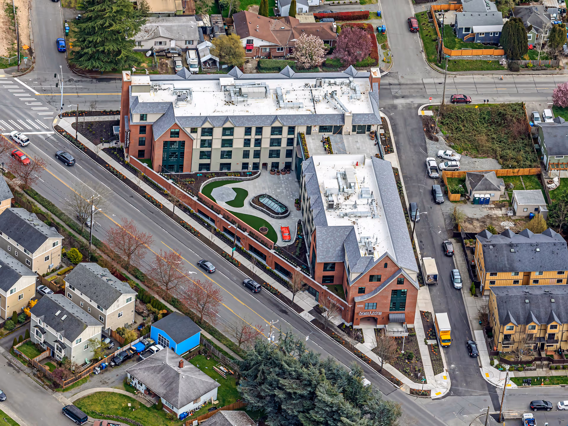 Aerial view of Aegis Living Greenwood, a multi-story senior living facility with a white roof and brick exterior, surrounded by residential houses and streets with cars.