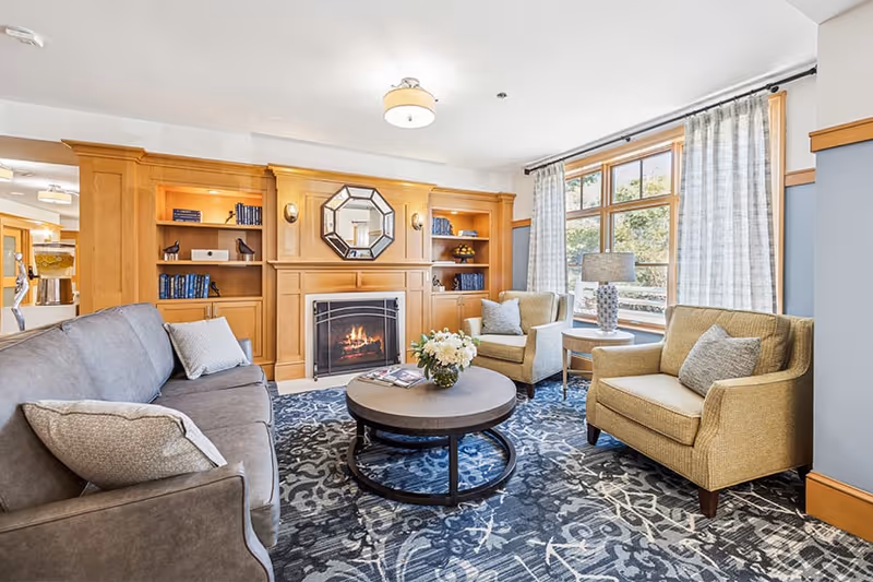 A cozy living room with a gray sofa and two beige armchairs arranged around a round coffee table with a flower vase on it. The room features a wooden built-in bookshelf and fireplace with a decorative mirror above it. Large windows with light curtains allow natural light to fill the space.
