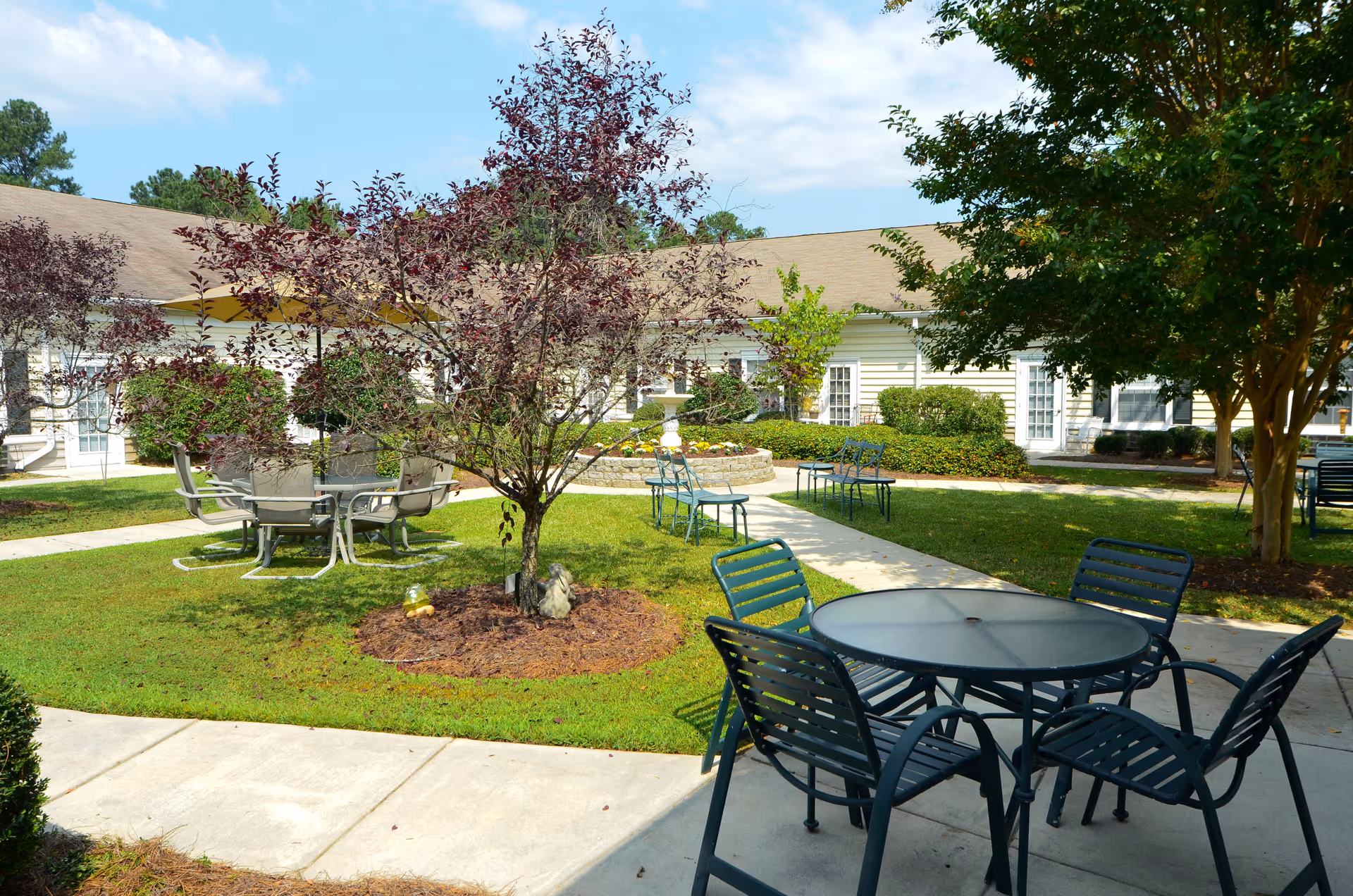 Outdoor courtyard area at Lancaster Grove Senior Living with several metal tables and chairs arranged on concrete pathways and grassy areas, surrounded by trees and shrubs, with a building in the background under a partly cloudy sky.