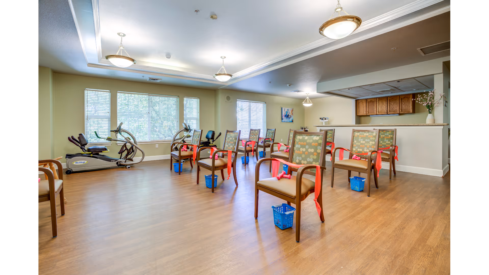 A spacious exercise room with wooden flooring and large windows letting in natural light. The room contains several chairs arranged in rows, each with red resistance bands tied to the arms and small blue baskets placed underneath. Two exercise bikes are positioned near the windows. The walls are painted light yellow, and there are ceiling lights and a small counter area with wooden cabinets in the background.