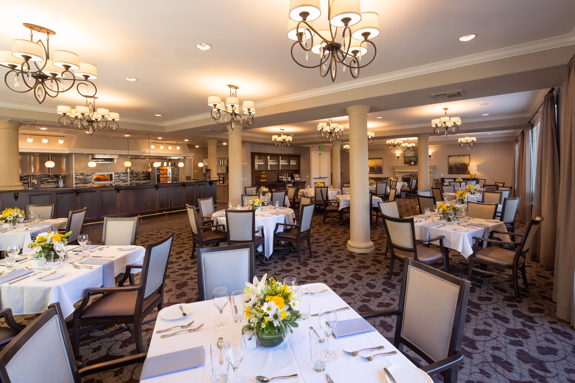 A spacious and elegant dining room with multiple round tables covered in white tablecloths, each set with glassware, silverware, napkins, and floral centerpieces. The room features several chandeliers hanging from the ceiling, large windows with curtains, and a serving area with a counter and kitchen appliances in the background.