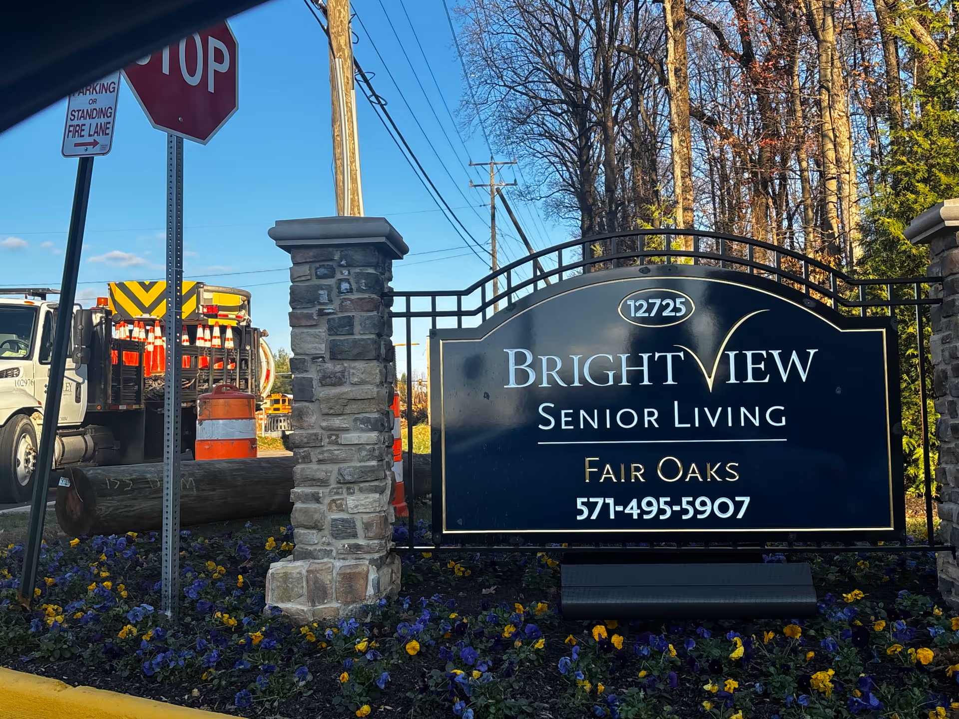 Outdoor entrance sign for Brightview Senior Living Fair Oaks with the address 12725 and phone number 571-495-5907, surrounded by flowers and stone pillars, with a stop sign and construction vehicles in the background.