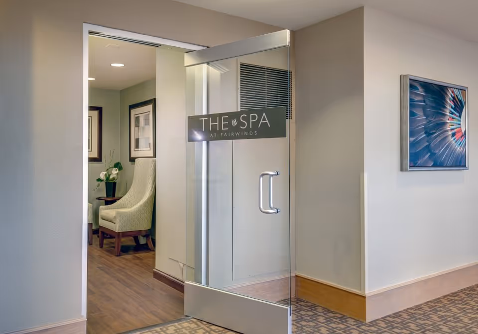 A glass door labeled "THE SPA AT FAIRWINDS" opens into a quiet interior seating area with upholstered chairs, artwork, and wood flooring.