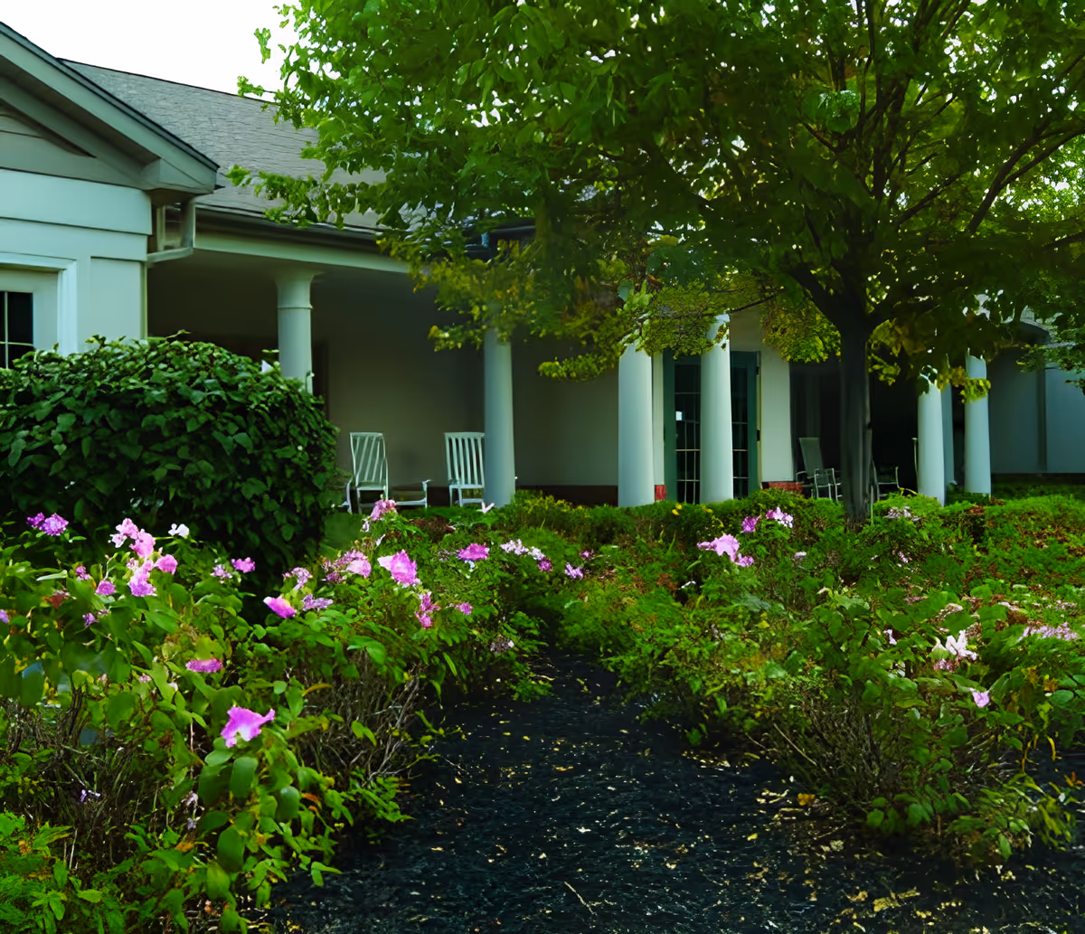 Front exterior of a senior living building with a covered porch, white columns, rocking chairs, and flower beds in the foreground.